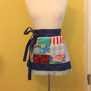 May include: A patchwork apron with a blue denim base, featuring a variety of colorful floral and geometric patterns. The apron has a tie closure and a lace trim.