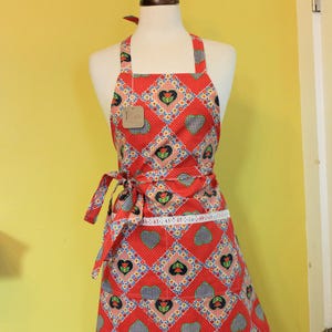 May include: A red apron with a repeating pattern of hearts, flowers, and geometric shapes. The apron has a white lace trim along the bottom and a tie at the waist. The apron is displayed on a mannequin.