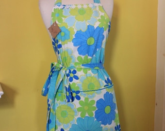 May include: A vintage-style apron with a floral pattern in shades of blue and green on a white background. The apron has a halter neck, a waist tie, and a pocket. The apron is displayed on a mannequin.