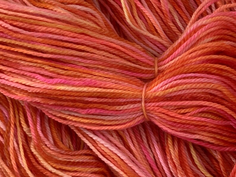 May include: Close-up of a skein of yarn in shades of orange, pink, and red. The yarn is soft and fluffy, and the colors are vibrant and rich.