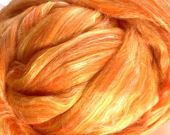 May include: A large, rounded bundle of unspun fiber roving featuring a vibrant blend of bright orange, golden yellow, and soft peach tones. The material has a lustrous, silky texture with long, flowing strands arranged in a dense, coiled formation.