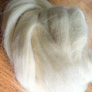 May include: A close-up of a pile of off-white wool roving. The fibers are long, soft, and slightly crimped, with a natural, textured appearance. The wool is arranged in a loose, flowing mass, ideal for fiber art projects.