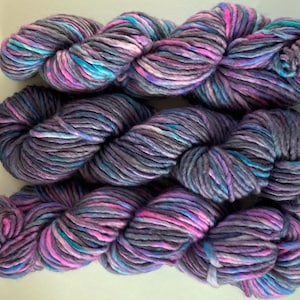 May include: A skein of yarn in a mix of purple, pink, blue, and gray colors. The yarn is thick and fluffy, and it is perfect for knitting or crocheting.