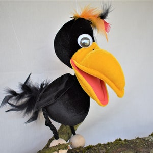 May include: A black crow puppet with a yellow beak and red mouth, perched on a branch. The crow has orange and black feathers on its head and black feathers on its body.