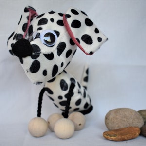 May include: A black and white spotted plush dog toy with googly eyes and pink ears. The dog is standing on four wooden balls.