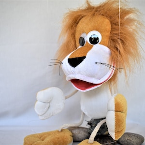 May include: A brown and white puppet lion with a red mouth and large white eyes. The lion has a long, brown mane and is wearing a white shirt. The puppet is sitting on a rock and is being held by a string.