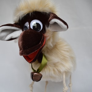 May include: A plush sheep puppet with a brown face, ears, and hat, and large, reflective eyes. The sheep has a fluffy, cream-colored body and a green ribbon with a small bell. The puppet is designed to be held by strings.