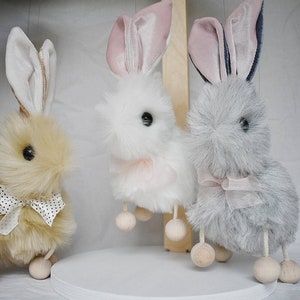 May include: Three plush bunny toys with wooden legs and pink ribbon accents. The bunnies are beige, white, and gray.