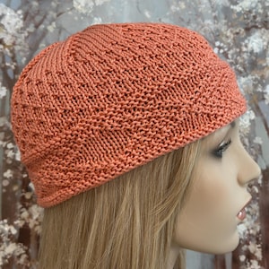 May include: A knitted coral-colored beanie hat. The hat is made of a textured knit fabric and has a rounded crown. The hat is shown on a mannequin with blonde hair. The background is a blurred floral painting.