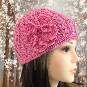 Puede incluir: Un gorro de punto rosa con una gran flor rosa en el lateral. El gorro está hecho con un delicado patrón de punto.