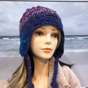 Puede incluir: Un gorro de punto azul, rosa y morado con orejeras. El gorro tiene un punto texturizado y un pompón en cada orejera.