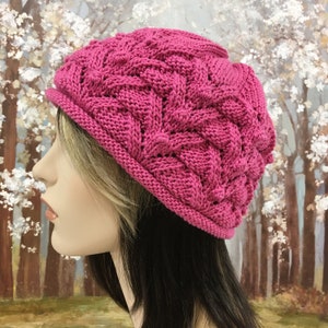 May include: A pink knitted hat with a cable knit pattern. The hat is worn on a mannequin with long brown hair.