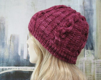 May include: A close-up of a burgundy knitted hat with a cable knit pattern. The hat is worn by a person with long blonde hair.