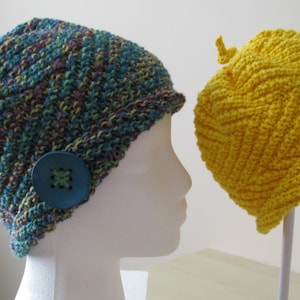 Op de afbeelding: Twee gehaakte mutsen met knopen en versieringen. De ene muts is blauw, groen en paars, de andere is geel. Beide mutsen worden getoond op witte mannequins.