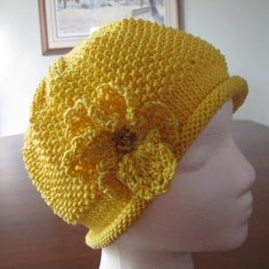 Può includere: Un cappello di lana giallo con un dettaglio floreale. Il cappello è realizzato in un filato morbido e testurizzato e ha un design semplice e classico.