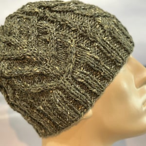 Puede incluir: Un gorro de punto verde con un patrón de trenza. El gorro está hecho con un hilo jaspeado que tiene un sutil brillo blanco.