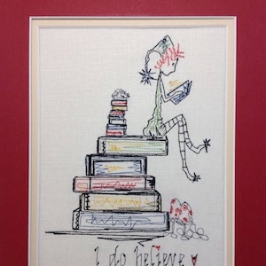 Puede incluir: Una obra bordada que representa a un elfo fantasioso sentado en una pila de libros, leyendo un libro. El elfo lleva un sombrero verde y tiene el pelo rojo. La obra está enmarcada e incluye el texto "I do believe".