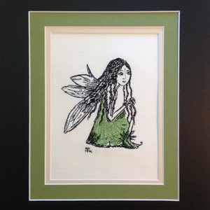 May include: A black and white embroidered image of a fairy with green wings and a green dress. The fairy is sitting with her head tilted down and her hands clasped in front of her. The image is framed in a green and white frame.