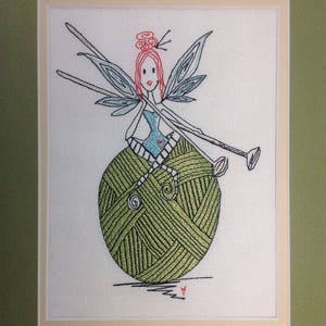 May include: Embroidered image of a fairy sitting on a ball of yarn, holding knitting needles. The fairy has red hair, blue wings, and a blue dress. The yarn is green.