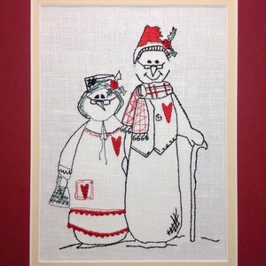 May include: Embroidered artwork of two snowmen, one wearing a red hat and a plaid scarf, the other wearing a red hat and a red heart on their chest. Both snowmen are outlined in black and have red accents.