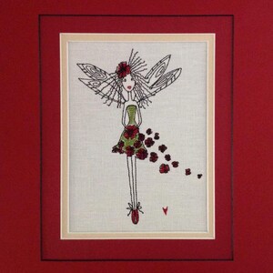 May include: Embroidered artwork featuring a whimsical fairy with red flowers in her hair and dress. The fairy has wings and is holding a bouquet of red flowers. The artwork is framed in a red mat with a cream-colored inner mat.