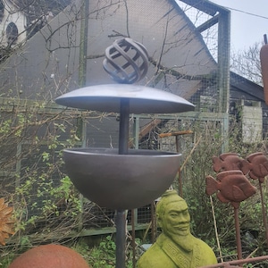 May include: A dark metal garden bird feeder with a spherical top and a bowl-shaped base. The feeder is mounted on a tall pole. The background includes a statue and metal fish decorations.