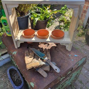 May include: A wooden plant stand with three terracotta pots. The stand has a rectangular top and angled side supports. Gardening gloves and a trowel are on a rusty metal container. The stand is made of light-colored wood.