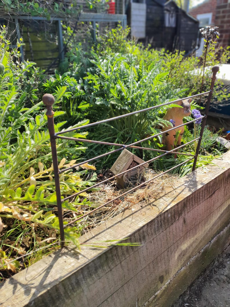 May include: A rustic metal garden fence with a decorative top, set in a raised wooden planter box. Lush green plants and a small terracotta animal statue are visible behind the fence. A small wooden insect house is also present.