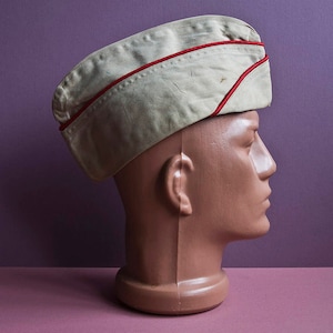 Puede incluir: Una gorra de estilo militar beige con ribete rojo. La gorra está en una cabeza de maniquí marrón.