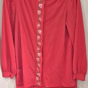 May include: A bright coral pink two-piece pajama set consisting of a long-sleeved button-down top and matching pants. The top features a vertical strip of white floral embroidery with green stems running down the center front placket.