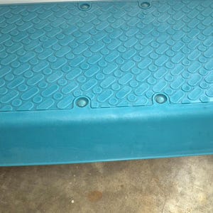 May include: A teal exercise step with a textured surface and pink feet. The step has the text "SI PI" on the top surface. This fitness equipment is designed for aerobic workouts and step exercises.