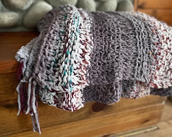 May include: A gray, white, and burgundy striped knitted throw blanket with fringe tassels. The blanket is folded and draped over a wooden surface.