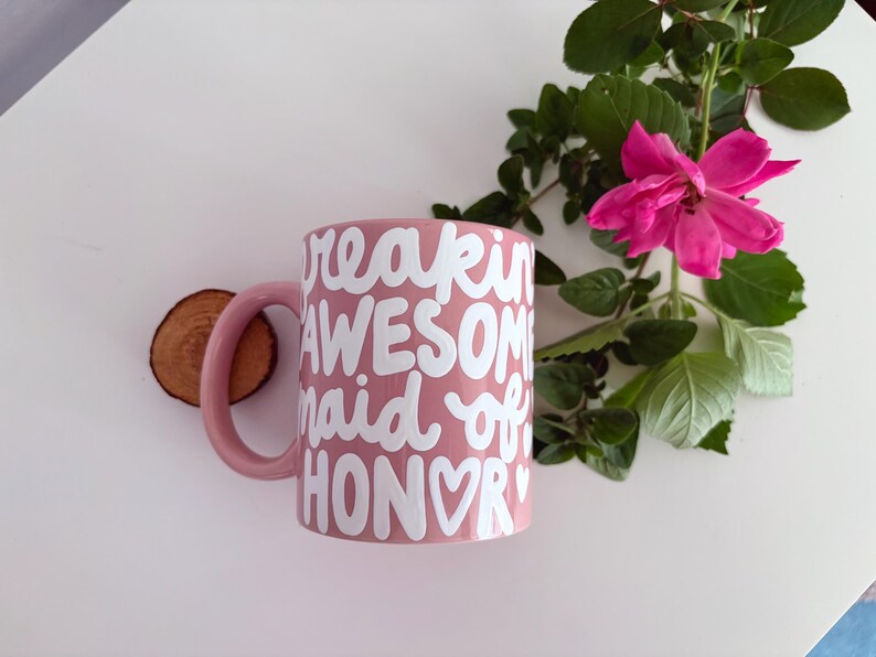 Puede incluir: Una taza de cer&aacute;mica rosa con las palabras blancas "freakin' AWESOME maid of HONOR". Una rosa rosa y vegetaci&oacute;n est&aacute;n al lado de la taza. Un peque&ntilde;o disco de madera tambi&eacute;n est&aacute; en la imagen.