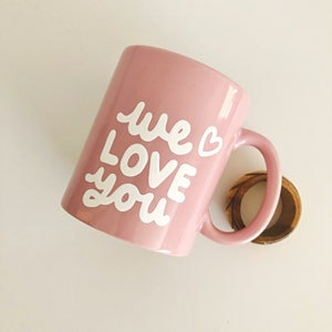 Puede incluir: Una taza de cer&aacute;mica rosa claro con las palabras "we LOVE you" en escritura blanca. Un peque&ntilde;o anillo de madera se encuentra cerca del asa. La taza es de color rosa claro y tiene un coraz&oacute;n junto a la palabra "we".