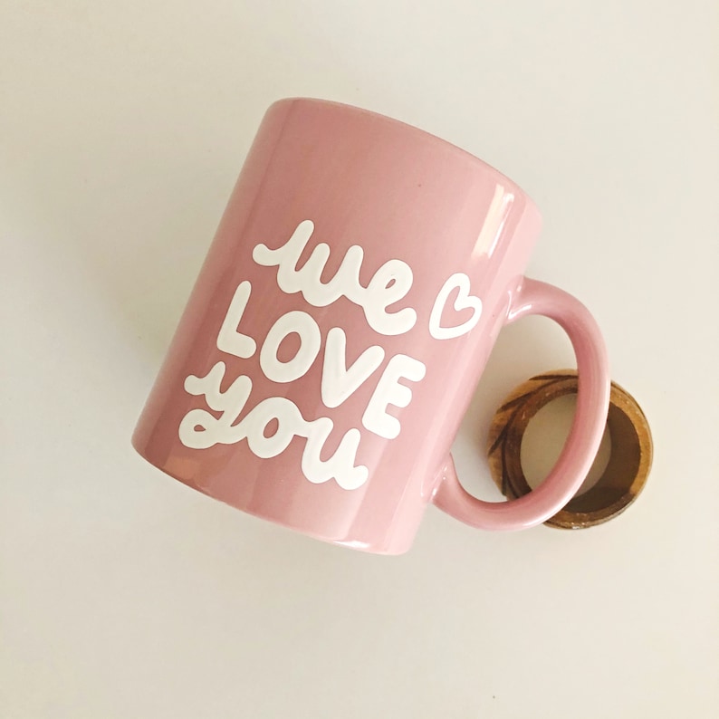 Puede incluir: Una taza de cer&aacute;mica rosa claro con las palabras "we LOVE you" en escritura blanca. Un peque&ntilde;o anillo de madera se encuentra cerca del asa. La taza es de color rosa claro y tiene un coraz&oacute;n junto a la palabra "we".