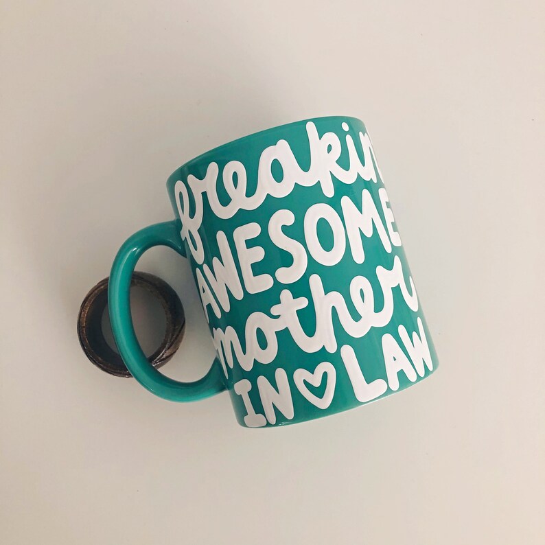 Puede incluir: Una taza de cer&aacute;mica turquesa con las palabras "freakin AWESOME mother IN LAW" en escritura blanca. La taza tiene un asa turquesa a juego y un anillo marr&oacute;n sujeto al asa.