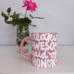 Puede incluir: Una taza de cer&aacute;mica rosa con las palabras "freakin' AWESOME maid of HONOR" en escritura blanca. La taza tiene un asa rosa a juego. Una rosa rosa y vegetaci&oacute;n est&aacute;n en un frasco de vidrio en el fondo.