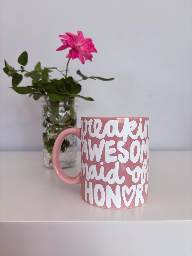 Puede incluir: Una taza de cer&aacute;mica rosa con las palabras "freakin' AWESOME maid of HONOR" en escritura blanca. La taza tiene un asa rosa a juego. Una rosa rosa y vegetaci&oacute;n est&aacute;n en un frasco de vidrio en el fondo.
