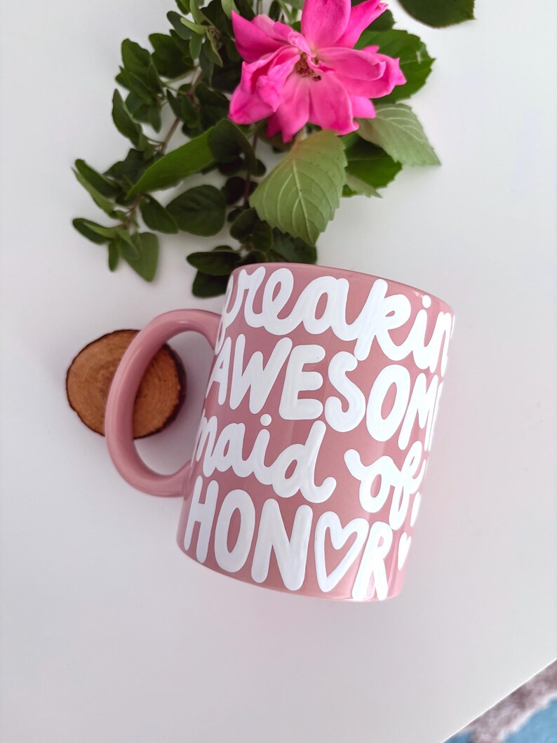 Puede incluir: Una taza de cer&aacute;mica rosa con las palabras "freakin AWESOME maid of HONOR" en letras blancas. La taza tiene un asa y est&aacute; junto a un peque&ntilde;o disco de madera, una rosa rosa y hojas verdes.