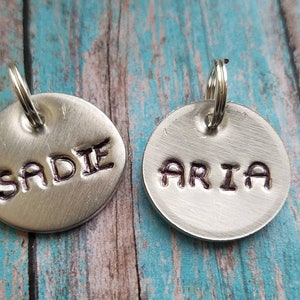 May include: Two silver dog tags with engraved names, "SADIE" and "ARIA", on a wooden background.