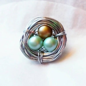 May include: A silver wire nest with three faux pearls inside. Two of the pearls are green and one is gold.