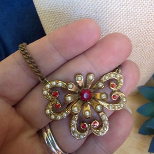 May include: A gold-toned metal necklace pendant shaped like a stylized flower or butterfly. The piece features a central red circular rhinestone, surrounded by small white faux pearls and additional red rhinestone accents set in a scrolling frame.