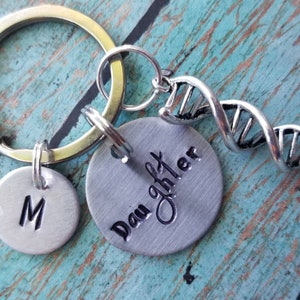Puede incluir: Llavero de plata con un dije de ADN, un dije redondo que dice "Hija", y un dije redondo con la letra "M".