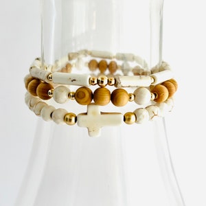 May include: Three stacked bracelets with white and brown beads and a gold cross charm.