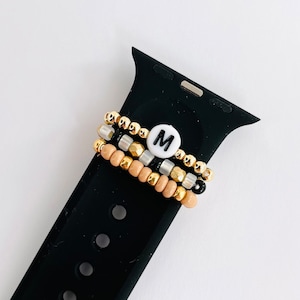 May include: Three beaded bracelets with a gold, silver, and black color scheme. The bracelets are stacked on a black Apple Watch band. The top bracelet has a white bead with the letter "M" on it.