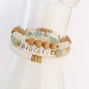 May include: A set of three bracelets featuring wooden beads, white beads, and light green beads. The middle bracelet has gold beads and white letter beads spelling out the word "PRAY".