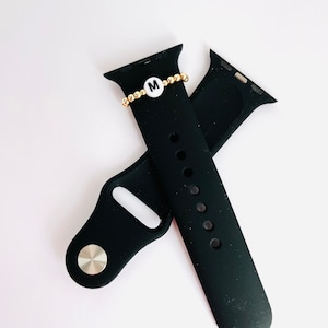 May include: Black silicone Apple Watch band with a gold beaded bracelet featuring a white charm with the letter 'M'.