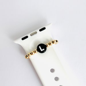 May include: A gold beaded Apple Watch band charm with a black circular bead featuring the white letter "L". The charm is attached to a white Apple Watch band. The beads are approximately 0.2 inches in diameter.