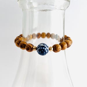 May include: A beaded bracelet featuring wooden beads with a striped pattern, interspersed with a blue and white patterned bead and gold-colored accents. The bracelet is displayed on a clear glass object.