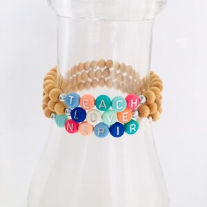 May include: A colorful beaded bracelet with the words "TEACH LOVE INSPIRE" spelled out in white letters on the beads. The bracelet is made of wooden beads and colorful beads.